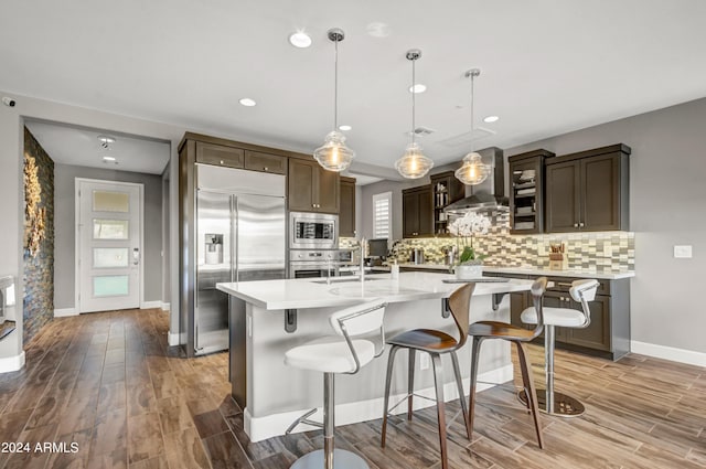 kitchen featuring a kitchen island with sink, light countertops, wall chimney range hood, and built in appliances