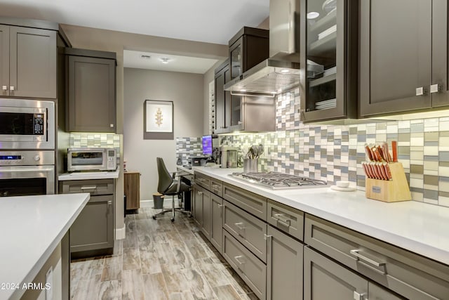 kitchen with tasteful backsplash, wall chimney exhaust hood, appliances with stainless steel finishes, glass insert cabinets, and light countertops