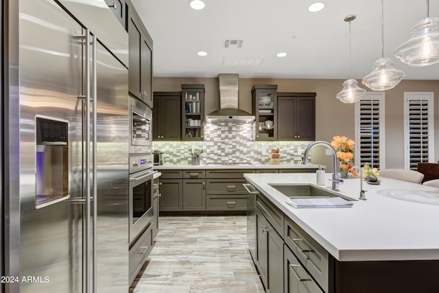 kitchen with light countertops, glass insert cabinets, wall chimney range hood, an island with sink, and built in appliances