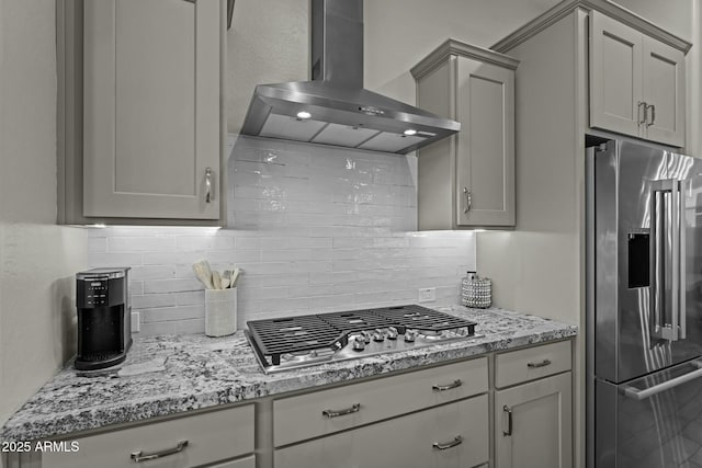 kitchen with light stone counters, gray cabinetry, stainless steel appliances, wall chimney exhaust hood, and tasteful backsplash