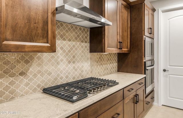 kitchen featuring light tile patterned floors, wall chimney range hood, light stone counters, stainless steel appliances, and backsplash