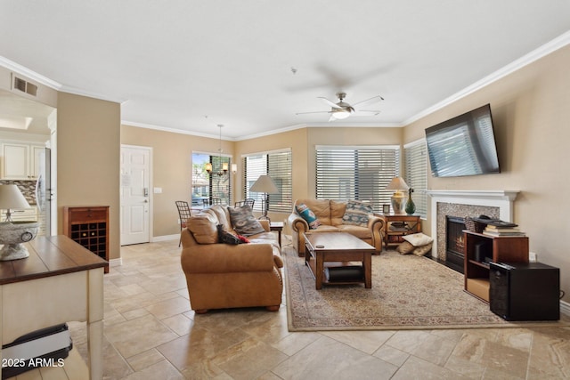 living room with a ceiling fan, a warm lit fireplace, baseboards, ornamental molding, and stone tile floors