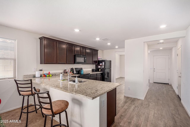 kitchen with recessed lighting, a sink, black appliances, a breakfast bar area, and dark brown cabinets