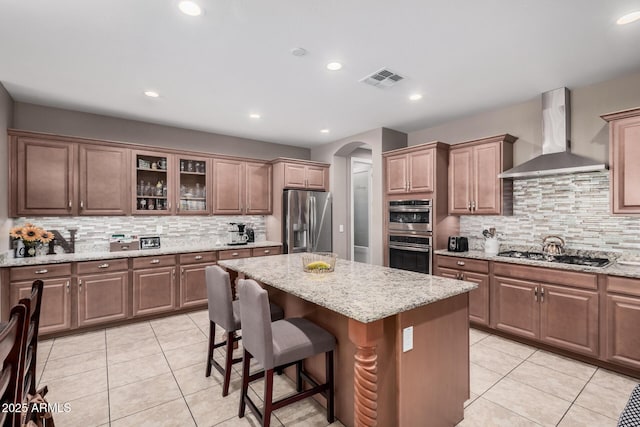 kitchen with arched walkways, a breakfast bar, wall chimney range hood, appliances with stainless steel finishes, and a center island