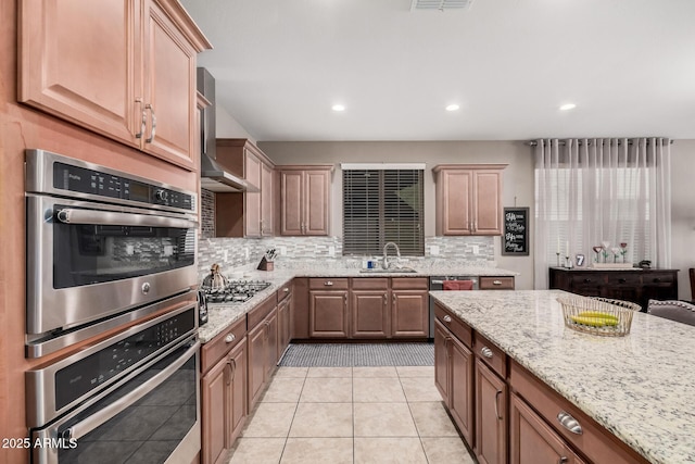 kitchen featuring light stone counters, decorative backsplash, appliances with stainless steel finishes, a sink, and wall chimney range hood