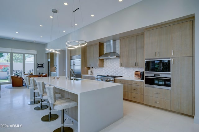 kitchen featuring a kitchen island with sink, wall chimney range hood, hanging light fixtures, appliances with stainless steel finishes, and light brown cabinetry