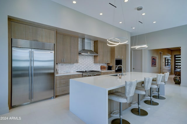 kitchen with wall chimney range hood, a center island with sink, sink, stainless steel appliances, and hanging light fixtures