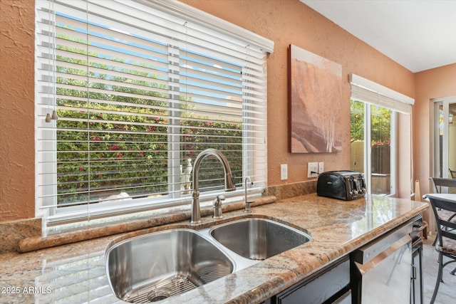 kitchen with a textured wall, a sink, light stone counters, and stainless steel dishwasher