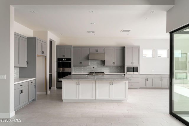 kitchen featuring double oven, gray cabinetry, tasteful backsplash, and an island with sink