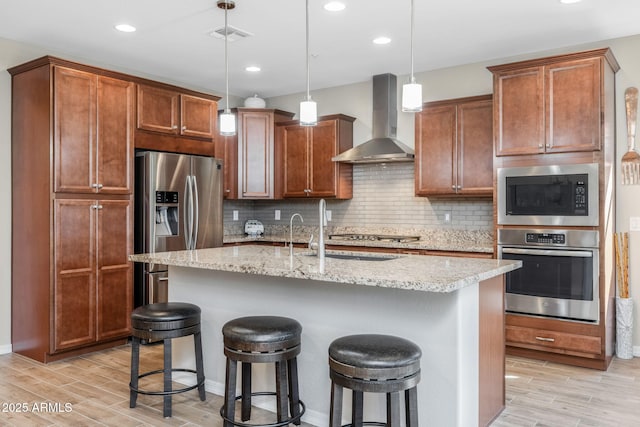 kitchen featuring wall chimney exhaust hood, appliances with stainless steel finishes, a kitchen island with sink, and pendant lighting