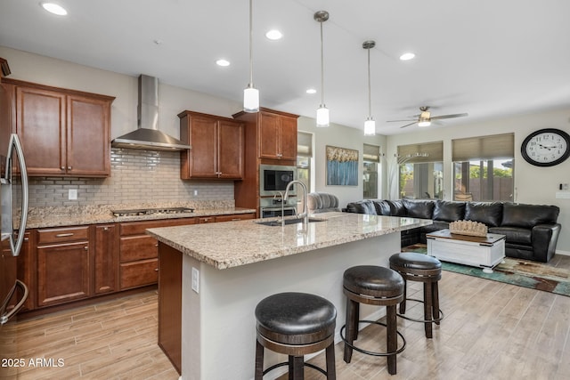 kitchen with sink, a kitchen breakfast bar, hanging light fixtures, a center island with sink, and wall chimney exhaust hood