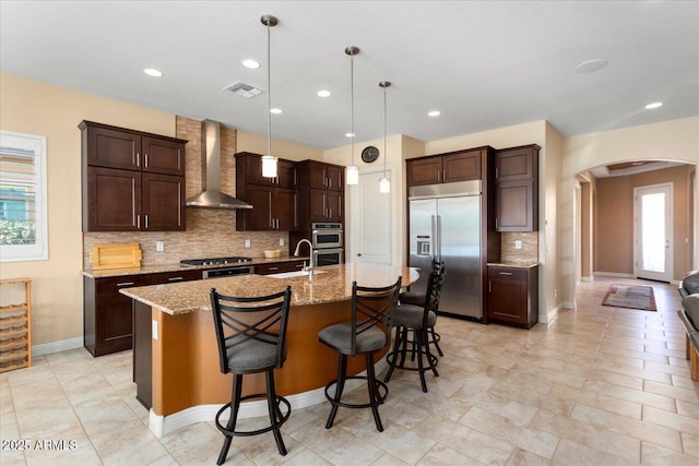 kitchen featuring pendant lighting, wall chimney exhaust hood, a kitchen bar, stainless steel appliances, and a center island with sink