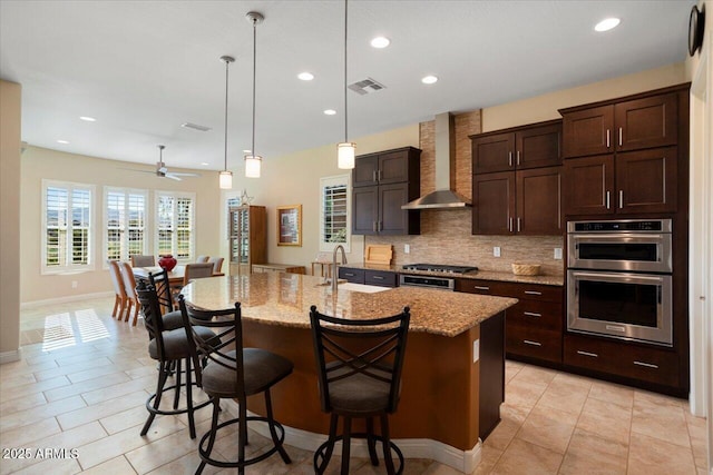 kitchen featuring wall chimney exhaust hood, sink, light stone counters, a kitchen island with sink, and a breakfast bar