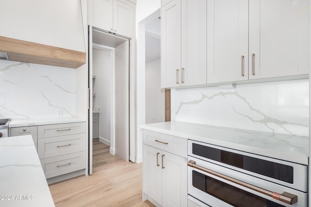 kitchen featuring light stone countertops, stainless steel oven, tasteful backsplash, light hardwood / wood-style flooring, and white cabinets