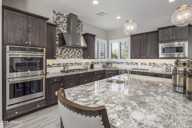 kitchen featuring light stone countertops, appliances with stainless steel finishes, decorative backsplash, wall chimney exhaust hood, and sink