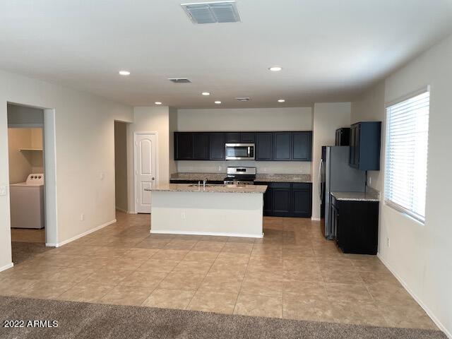 kitchen with an island with sink, light tile floors, light stone countertops, appliances with stainless steel finishes, and washer / dryer