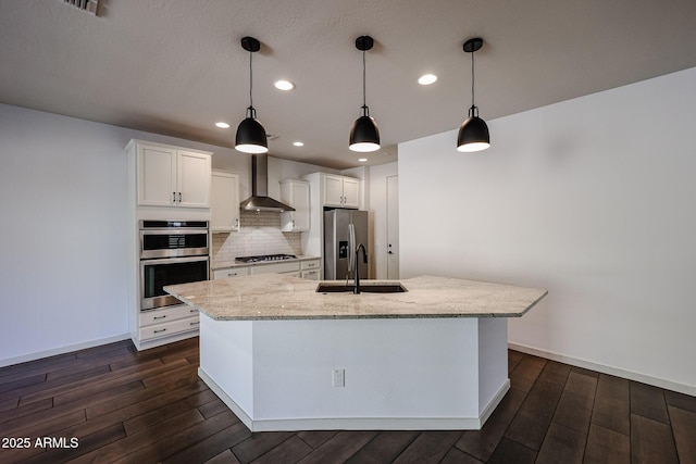 kitchen featuring appliances with stainless steel finishes, decorative light fixtures, white cabinetry, an island with sink, and wall chimney exhaust hood