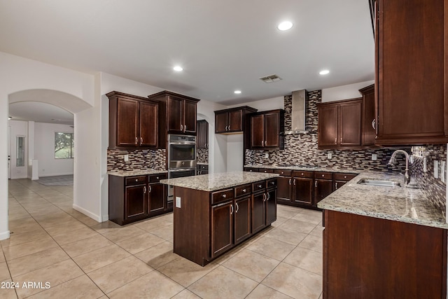 kitchen featuring double oven, light stone counters, arched walkways, wall chimney exhaust hood, and a sink