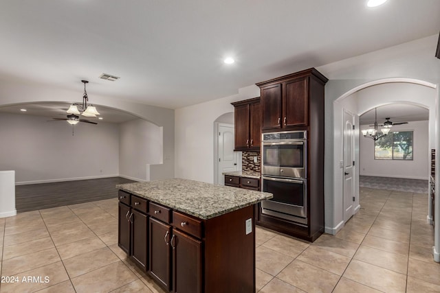 kitchen with light tile patterned floors, stainless steel double oven, arched walkways, ceiling fan with notable chandelier, and tasteful backsplash