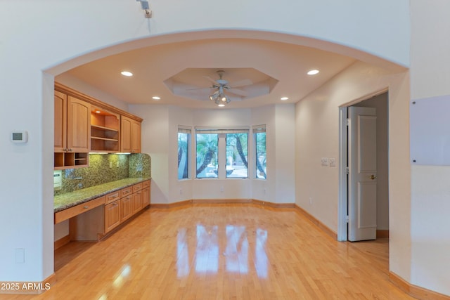 interior space with light wood-type flooring, a ceiling fan, a tray ceiling, recessed lighting, and arched walkways