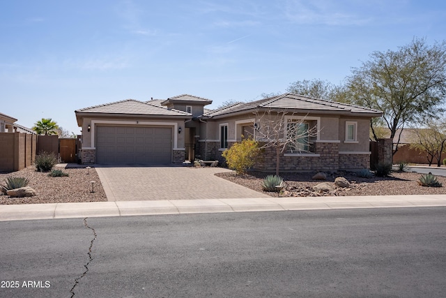 prairie-style home featuring a garage, fence, stone siding, decorative driveway, and stucco siding