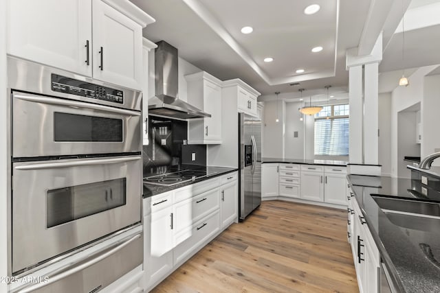 kitchen featuring a tray ceiling, light wood-style flooring, appliances with stainless steel finishes, white cabinetry, and wall chimney exhaust hood