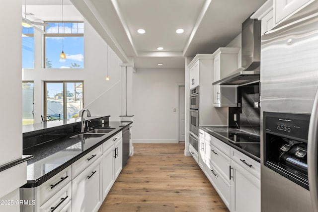 kitchen featuring stainless steel appliances, white cabinets, a sink, wall chimney range hood, and light wood-type flooring