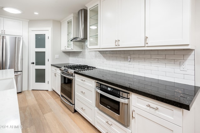 kitchen with white cabinetry, stainless steel appliances, light wood-type flooring, dark stone counters, and wall chimney range hood