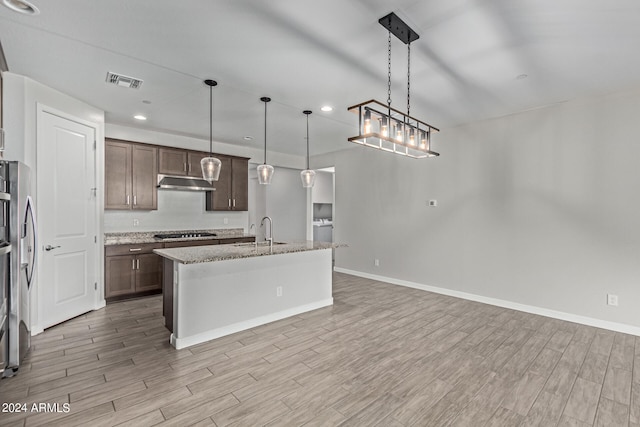 kitchen featuring an island with sink, light stone counters, decorative light fixtures, sink, and light hardwood / wood-style flooring