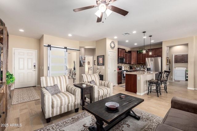 living area featuring visible vents, ceiling fan, a barn door, light wood-style floors, and washer / clothes dryer