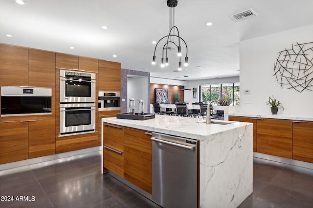 kitchen featuring appliances with stainless steel finishes, decorative light fixtures, sink, light stone counters, and a center island with sink