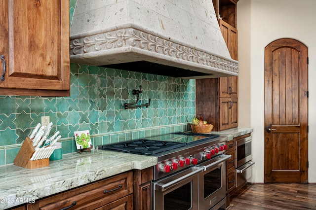 kitchen with dark wood-type flooring, backsplash, light stone counters, range with two ovens, and premium range hood