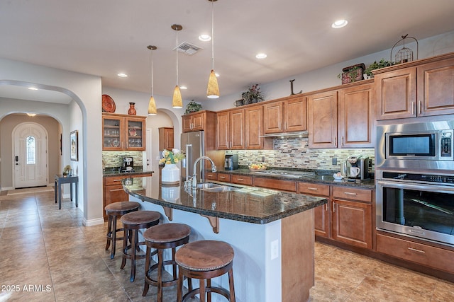 kitchen featuring a center island with sink, appliances with stainless steel finishes, pendant lighting, dark stone counters, and decorative backsplash