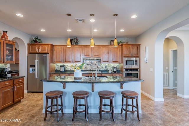 kitchen with pendant lighting, appliances with stainless steel finishes, a kitchen island with sink, decorative backsplash, and dark stone counters