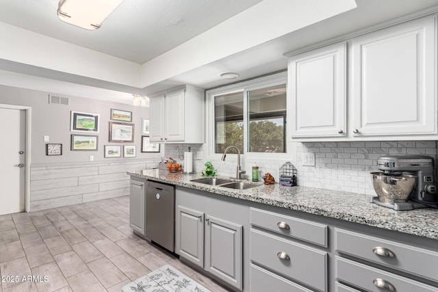 kitchen with a sink, light stone counters, stainless steel dishwasher, gray cabinetry, and wainscoting