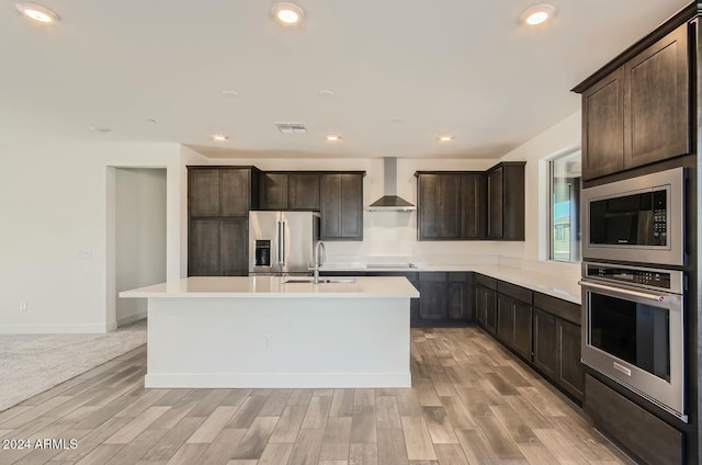 kitchen featuring stainless steel appliances, a kitchen island with sink, sink, wall chimney range hood, and light hardwood / wood-style flooring