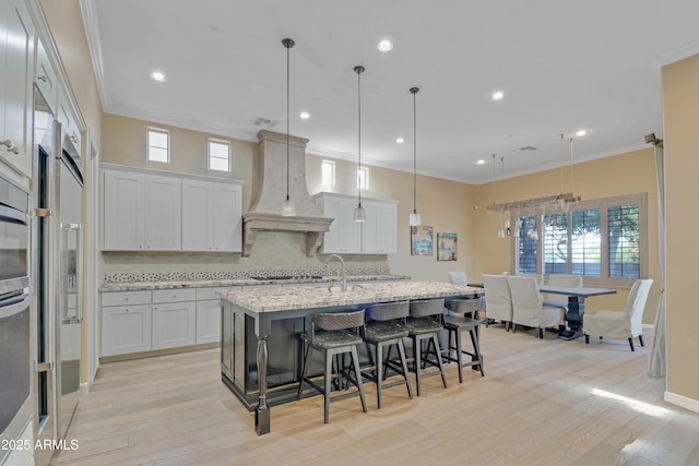 kitchen featuring white cabinetry, an island with sink, decorative light fixtures, and custom range hood