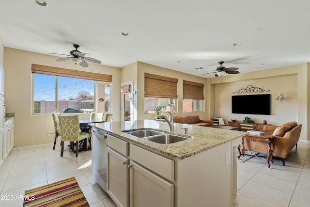 kitchen with light tile patterned flooring, dishwasher, sink, light stone counters, and a center island with sink