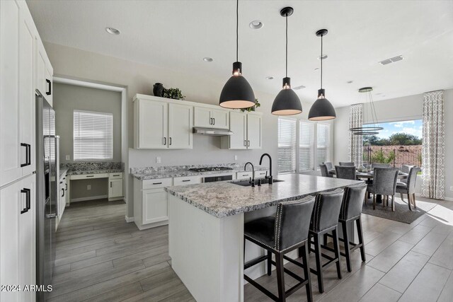 kitchen with white cabinets, wood-type flooring, decorative light fixtures, a center island with sink, and sink