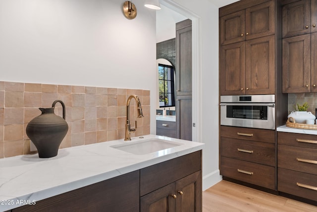 kitchen featuring oven, dark brown cabinets, a sink, light wood finished floors, and decorative backsplash