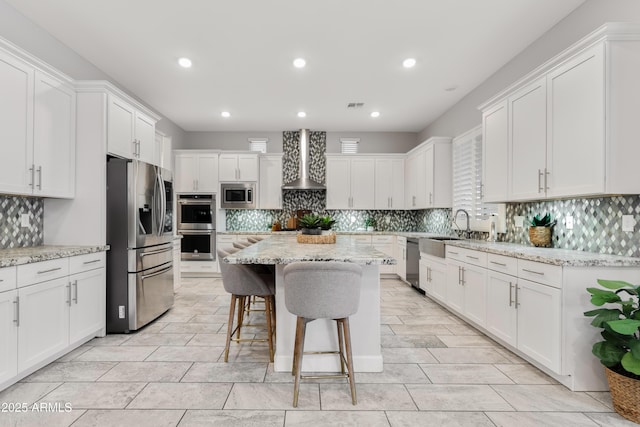 kitchen with a center island, appliances with stainless steel finishes, white cabinetry, wall chimney range hood, and light stone countertops