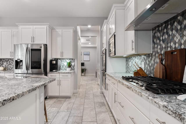 kitchen featuring appliances with stainless steel finishes, light stone countertops, wall chimney range hood, white cabinetry, and backsplash