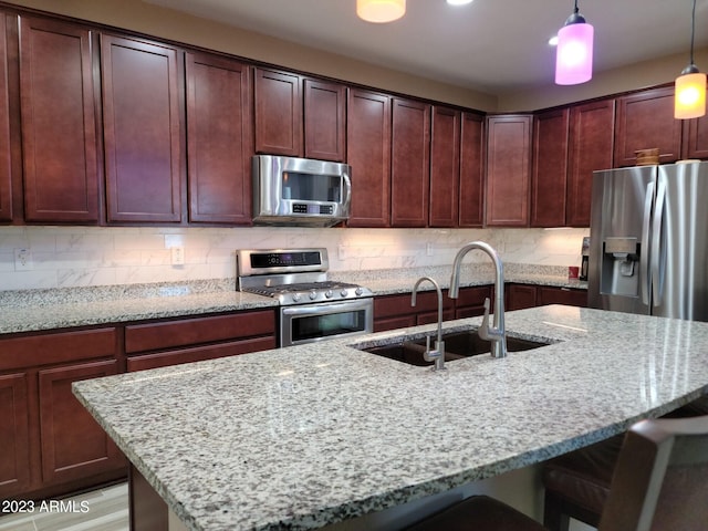 kitchen featuring hanging light fixtures, backsplash, stainless steel appliances, a kitchen island with sink, and light stone counters