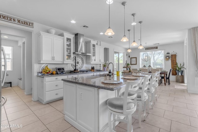 kitchen with wall chimney exhaust hood, pendant lighting, light tile flooring, white cabinetry, and a kitchen island with sink