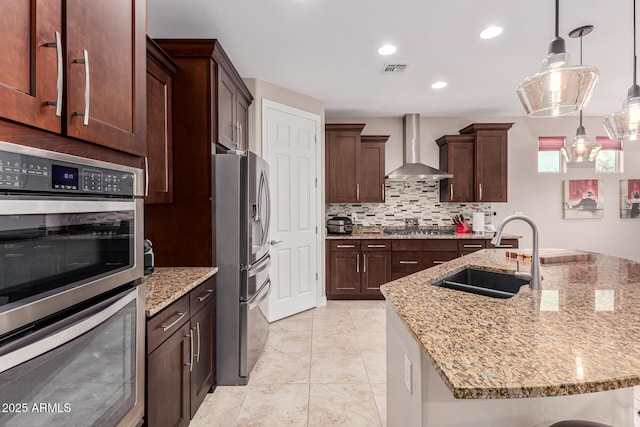 kitchen with light stone counters, stainless steel appliances, sink, wall chimney range hood, and pendant lighting