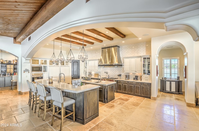 kitchen featuring backsplash, appliances with stainless steel finishes, an island with sink, beamed ceiling, and wall chimney exhaust hood