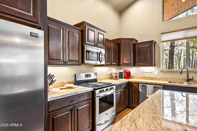 kitchen with light stone countertops, a sink, stainless steel appliances, dark brown cabinetry, and a towering ceiling