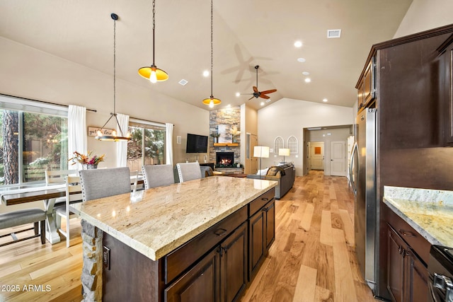 kitchen with light wood finished floors, a stone fireplace, dark brown cabinets, decorative light fixtures, and a center island