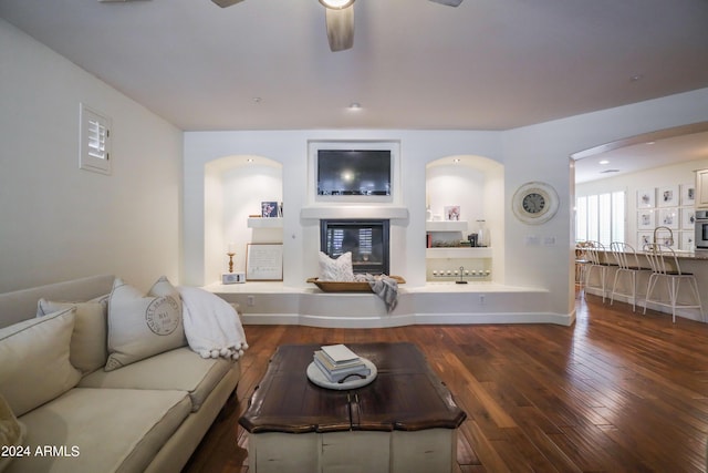 living room with dark wood-type flooring, ceiling fan, and built in shelves