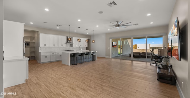 kitchen with wall chimney exhaust hood, white cabinetry, an island with sink, hanging light fixtures, and ceiling fan